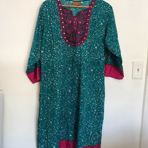 Puede incluir: Una t&uacute;nica azul turquesa con un cuello y ribetes bordados en rosa y negro. La t&uacute;nica tiene una peque&ntilde;a etiqueta blanca que dice "Folk Wear".