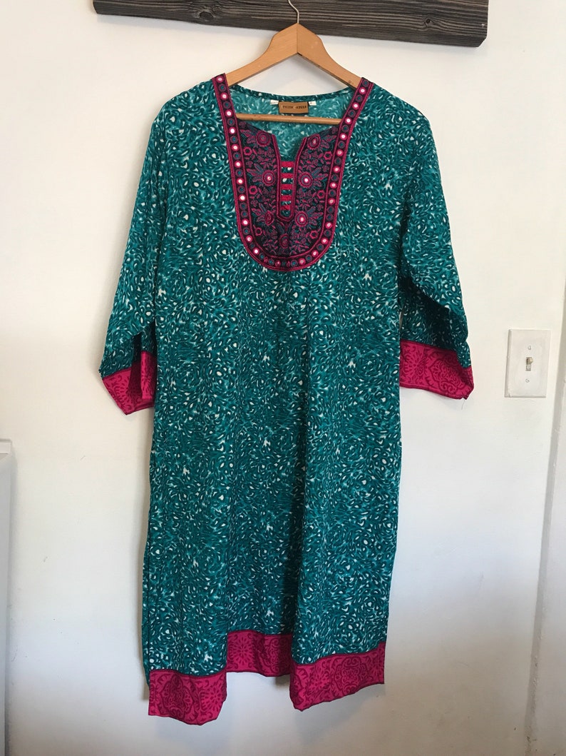 Puede incluir: Una t&uacute;nica azul turquesa con un cuello y ribetes bordados en rosa y negro. La t&uacute;nica tiene una peque&ntilde;a etiqueta blanca que dice "Folk Wear".