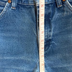 May include: Blue denim shorts with a high-waisted design, featuring a button fly, belt loops, and frayed raw-edge hems. A white measuring tape is placed vertically over the center, showing a rise measurement of approximately 12.5 inches.