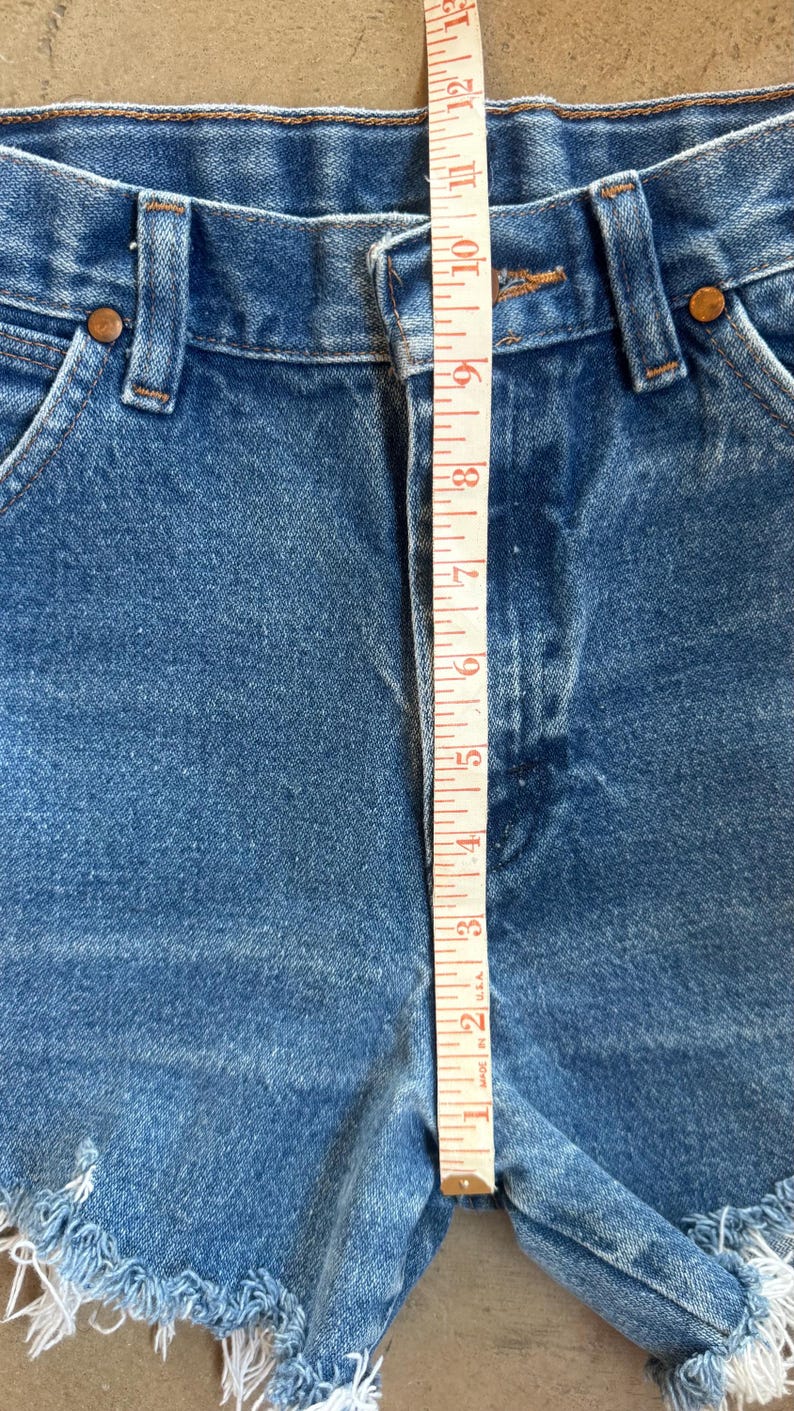 May include: Blue denim shorts with a high-waisted design, featuring a button fly, belt loops, and frayed raw-edge hems. A white measuring tape is placed vertically over the center, showing a rise measurement of approximately 12.5 inches.