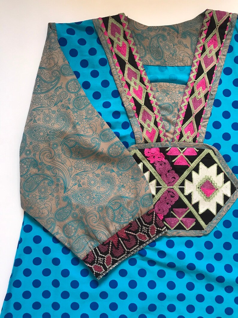 以下が含まれることがあります： ピンクと黒の刺繍が施されたネックラインと袖のある、青と茶色の模様のチュニック。チュニックは、前面と背面に水玉模様があります。