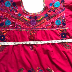 May include: A bright pink embroidered blouse with colourful floral designs. The blouse has a round neckline and short sleeves. A measuring tape is placed across the bottom of the blouse, showing a measurement of 63.5 cm.