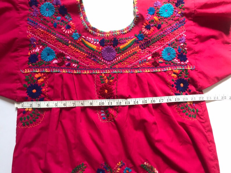 May include: A bright pink embroidered blouse with colourful floral designs. The blouse has a round neckline and short sleeves. A measuring tape is placed across the bottom of the blouse, showing a measurement of 63.5 cm.