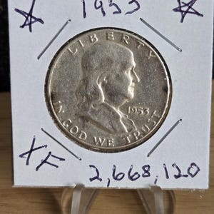 May include: A 1953 silver coin featuring a profile of Benjamin Franklin, with the inscriptions "LIBERTY" and "IN GOD WE TRUST." The coin is displayed in a white holder with handwritten markings including "1953," "XF," and "2,668,120."