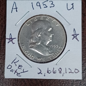 May include: A silver US half dollar coin with the year 1953 on the front. The coin is in a plastic coin holder with the text "A 1953 U" and "Key Date 2,668,120" written on it.