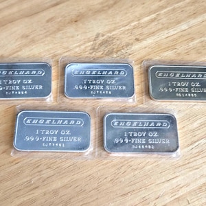 May include: Five silver bars, each encased in clear plastic. The bars are rectangular with rounded corners and feature the text "ENGELHARD", "1 TROY OZ.", and ".999 FINE SILVER". The bars are displayed on a wooden surface.