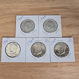 May include: Five silver Kennedy half dollars in protective paper holders. The coins are dated 1965, 1966, 1967, 1968, and 1969. The coins are displayed on a wooden surface.