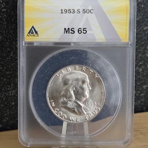 May include: A silver 1953-S 50C coin, graded MS 65, encased in a clear protective holder. The coin features a profile of a man and the inscription "IN GOD WE TRUST". The holder displays the text "1953-S 50C MS 65" and the ANACS logo.