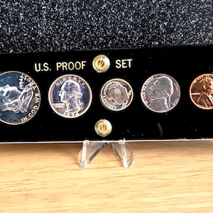 May include: A 1954 U.S. Proof Set displayed in a black case. The set includes a silver half dollar, quarter, dime, nickel, and a copper penny. The case is labeled "U.S. PROOF SET" in white text and is mounted on a clear stand.