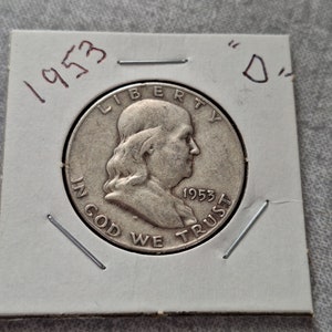 May include: A silver 1953 United States half dollar coin with the inscription "LIBERTY" above a profile of Benjamin Franklin and "IN GOD WE TRUST" below. The coin also has the year "1953" inscribed on the right side.