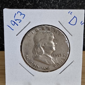 May include: A silver 1953 Franklin half dollar coin in a protective holder. The coin features a profile of Benjamin Franklin and the words "IN GOD WE TRUST" and "LIBERTY". The holder has the year "1953" and the letter "D" written on it.