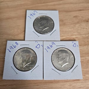 May include: Three silver Kennedy half dollars in protective paper holders. The coins are dated 1967, 1968, and 1969, with the 1969 coin marked with a "D". The coins are displayed on a light brown wooden surface.