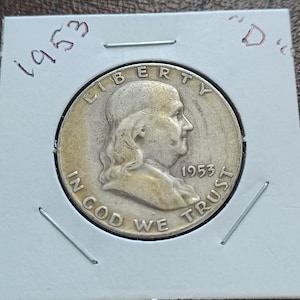 May include: A silver US half dollar coin with the profile of Franklin D. Roosevelt on the obverse. The coin is dated 1953 and has the inscription "LIBERTY" above the profile and "IN GOD WE TRUST" below. The coin is in a coin holder with the year 1953 and the letter "D" written on it.