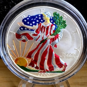 May include: A silver coin encased in a clear protective case. The coin features a colorful depiction of a robed figure holding a branch, with a waving American flag in the background. The coin is dated 2000 and includes the text "IN GOD WE TRUST".
