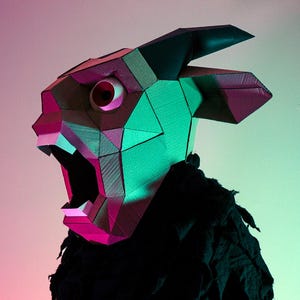 Könnte beinhalten: Eine geometrische Maske in Form eines gehörnten Wesens, mit offenem Mund und einem einzelnen sichtbaren Auge. Die Maske ist hauptsächlich braun, mit rosa und grünen Akzenten, vor einem rosa-grünen Farbverlauf. Die Maske wird über schwarzer Kleidung getragen.