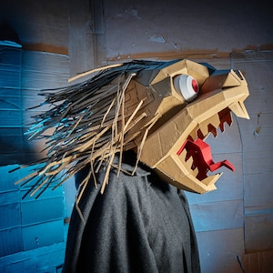 Op de afbeelding: Een handgemaakt drakenmasker gemaakt van karton. Het masker heeft een gedetailleerd ontwerp met een wijd open mond, een rode tong en een groot wit en rood oog. De drakenkop is bruin met bruine accenten en lang, gefranjerd haar.