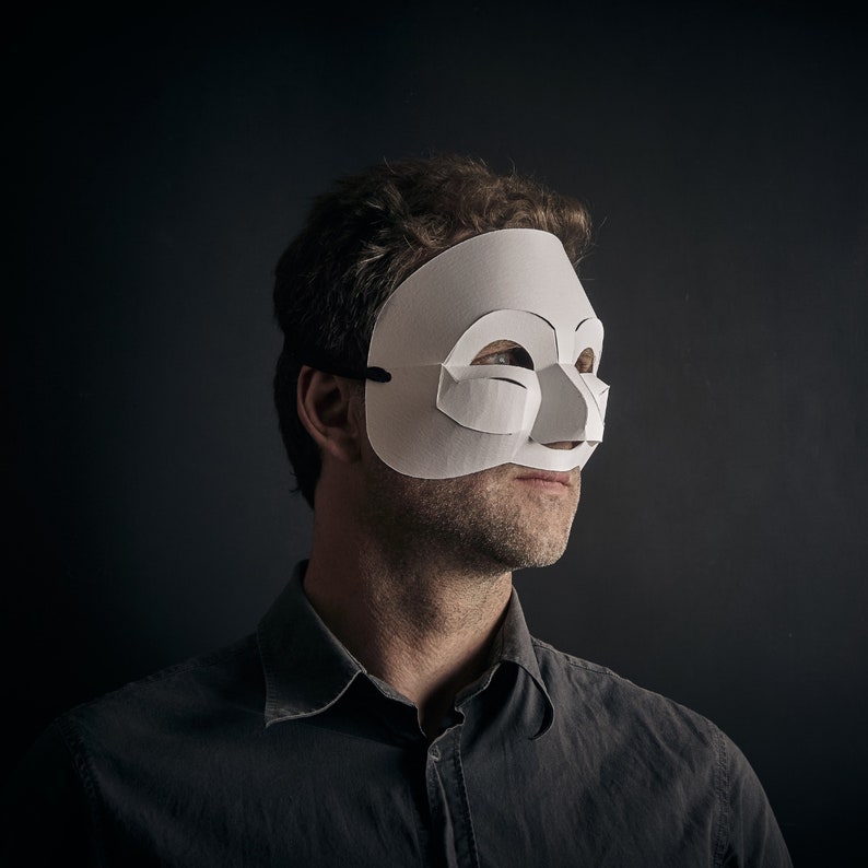 May include: A person wearing a white paper mask with a simple design. The mask covers the eyes and nose, and the person is looking to the right. The person is wearing a dark shirt.