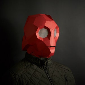 Puede incluir: Una persona lleva una máscara de papel maché roja con un diseño geométrico y protecciones para los ojos de malla. La máscara cubre la cara y la cabeza de la persona. La persona lleva una chaqueta verde oscura.