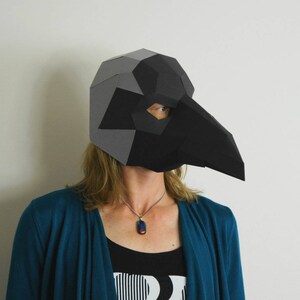 May include: A person wearing a black and gray paper mache mask shaped like a bird's head. The mask has a long beak and covers the person's eyes. The person is wearing a teal blue cardigan and a black tank top with a white and black striped pattern. The person is wearing a silver necklace with a dark blue and red pendant.
