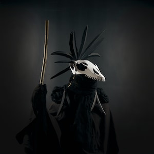 May include: A person wearing a black hooded robe and a white skull mask with a spiky black headdress. The person is holding a wooden staff.