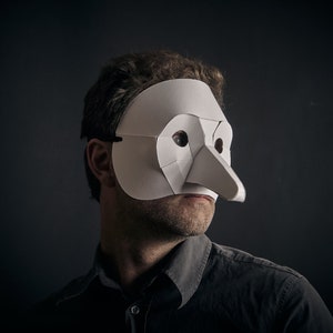 May include: A person wearing a white paper mask with a long nose. The mask is covering the person's eyes and nose. The person is wearing a dark shirt.