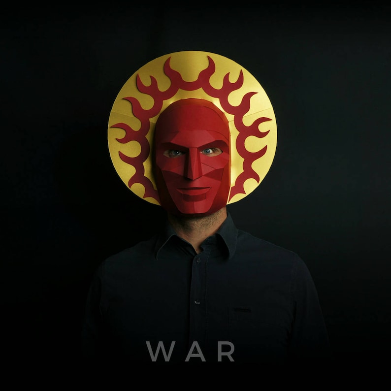 May include: A person wearing a red paper mask with a gold and red halo. The mask has a fierce expression and the halo has flames. The person is wearing a black shirt and the word "WAR" is printed below the image.