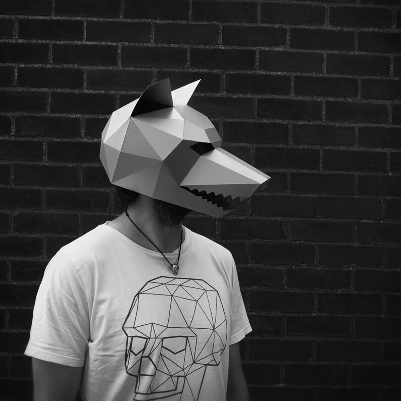 以下が含まれることがあります： 幾何学模様のグレーの紙粘土製のオオカミのマスクをつけた人。マスクは顔全体を覆っており、鋭く尖った鼻先をしています。その人は、黒い幾何学模様の骸骨のデザインが施された白いTシャツを着ています。