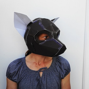 May include: A person wearing a black and gray paper mache mask with large ears. The mask is shaped like a dog's head and has a geometric design.