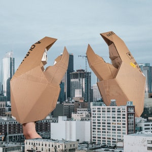 Puede incluir: Dos grandes manos de cartón, una con la palabra "NEER" impresa, están colocadas frente a un horizonte urbano. Las manos están hechas de cartón marrón y parecen estar extendiéndose.