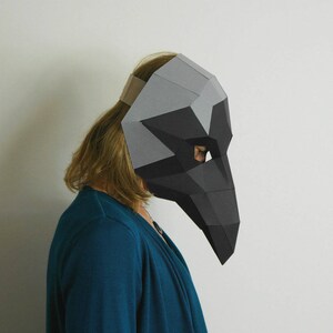 May include: A person wearing a black and gray paper mache mask with a long beak. The mask is covering the person's face and is held in place by a tan headband.