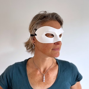 May include: A white masquerade mask with a black elastic strap. The mask has a curved shape and eye holes. The person wearing the mask is looking to the right.