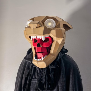 May include: A low-poly papercraft mask shaped like a tan panther head with white circular eyes and an open mouth revealing a red skull structure with white teeth, worn over a black hooded garment with a textured fabric collar.