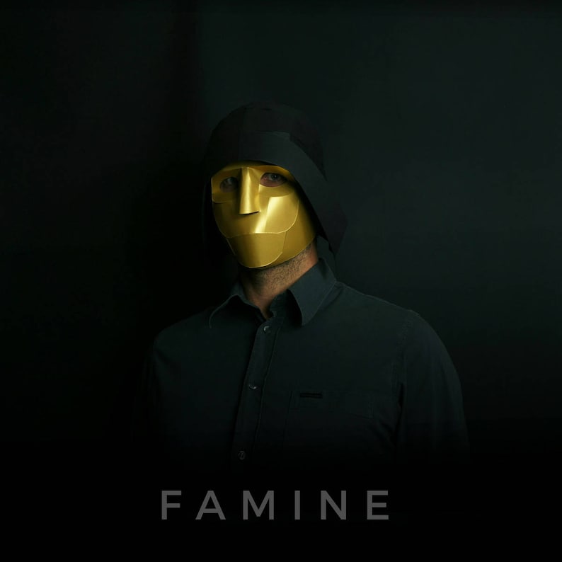 May include: A person wearing a black hooded garment and a gold mask with the word "FAMINE" printed below.