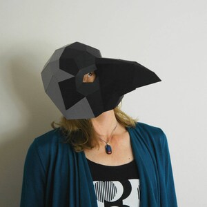 May include: A person wearing a black and gray paper mache mask shaped like a bird's head. The mask has a long beak and eye holes. The person is wearing a teal blue cardigan and a black tank top with a white and black graphic design.