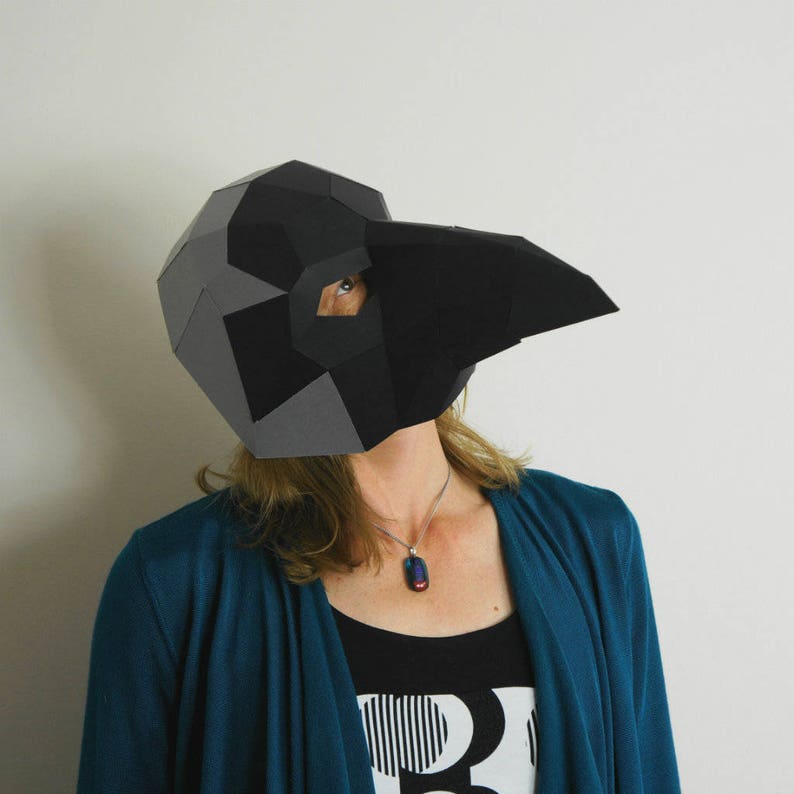 May include: A person wearing a black and gray paper mache mask shaped like a bird's head. The mask has a long beak and eye holes. The person is wearing a teal blue cardigan and a black tank top with a white and black graphic design.