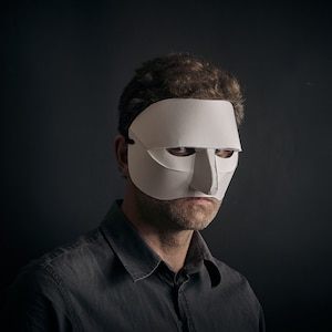 May include: A man wearing a white paper mask with a large, prominent nose. The mask covers his eyes and forehead. He is looking directly at the camera with a serious expression.