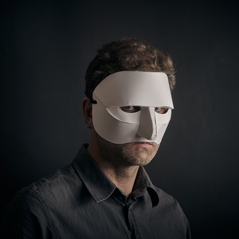May include: A man wearing a white paper mask with a large, prominent nose. The mask covers his eyes and forehead. He is looking directly at the camera with a serious expression.
