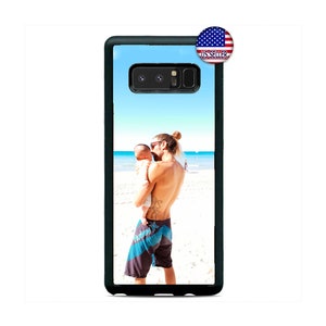 May include: A black phone case with a photo of a man holding a baby on a beach. The photo shows a blue sky, white sand, and blue water.
