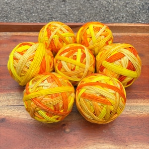 May include: Seven balls of yarn in shades of yellow and orange are arranged on a wooden surface. The yarn is wound in a crisscross pattern, creating a textured appearance. The color palette is warm and inviting.