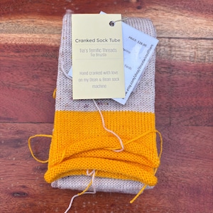 May include: A rolled sock tube in beige and yellow. The sock tube has a tag that reads "Cranked Sock Tube" and "Tia's Terrific Threads". The tag also states the socks are hand cranked with love on a Dean & Bean sock machine.