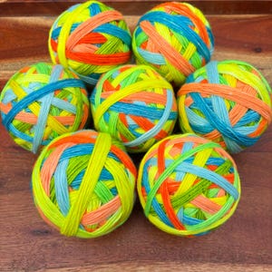 May include: Seven spherical balls of yarn arranged in a cluster, each featuring a vibrant, multi-colored pattern of neon yellow, bright orange, sky blue, and lime green strands wound tightly together in a crisscross, textured design.