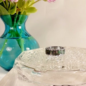 May include: A silver-toned metal ring featuring a band inlaid with rectangular segments of iridescent dark green and black abalone shell. The ring has a polished metallic finish and a flat profile, showcasing a repeating pattern of natural shell pieces.