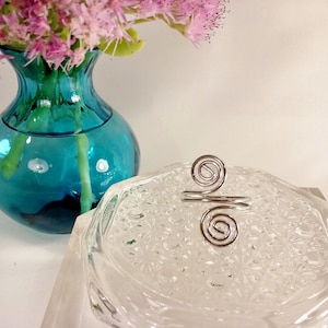 May include: A silver-colored ring with a double spiral design. The ring sits on a clear, textured glass surface. A turquoise vase with pink flowers is in the background.