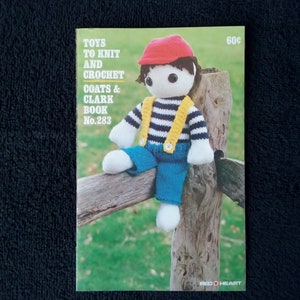 May include: A vintage book cover titled "Toys to Knit and Crochet" featuring a knitted doll wearing a red cap, blue pants, and a striped shirt with yellow suspenders. The book is from Coats & Clark, Book No. 283.