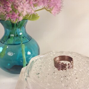 May include: A copper-colored ring with an openwork design sits on a clear, textured glass dish. The ring features a series of connected loops. A turquoise vase with pink flowers in the background adds a pop of color to the composition.