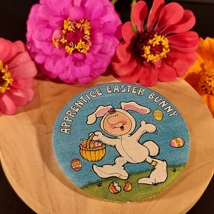 May include: A round, blue button with the text "Apprentice Easter Bunny" and a cartoon bunny carrying a basket of eggs. The button is surrounded by pink and orange flowers.