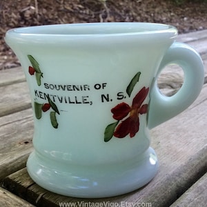 May include: A pale blue milk glass mug featuring a floral design with red petals and green leaves. The text "SOUVENIR OF KENTVILLE, N. S." is printed in black on the front. The mug has a wide rim, a flared base, and a rounded side handle.