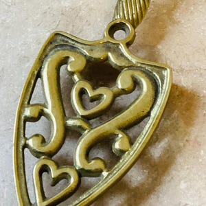 May include: A vintage brass trivet with a shield shape and a handle. The trivet features an openwork design with heart shapes and decorative swirls. The handle has a ribbed texture. The trivet is a warm gold color.