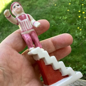 May include: A small ceramic figurine of a person wearing a pink and white striped outfit, standing on a white staircase structure with a red side panel. The figure has a painted face with blue eyes and is posed with one arm raised in a waving motion.