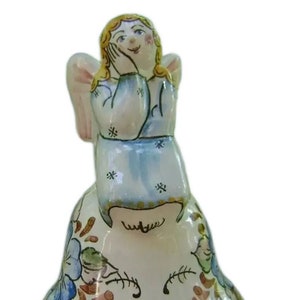 May include: A ceramic angel bell with a blue and white dress, yellow hair, and pink wings. The bell is decorated with floral designs in shades of brown, blue, and green. The bell has a yellow rim.